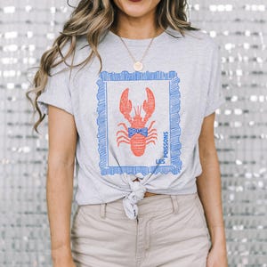 May include: A light gray t-shirt featuring a red lobster design within a blue frame. The lobster has a blue bow tie and the text "LES POISSONS" is printed on the side. The shirt is tied at the waist.