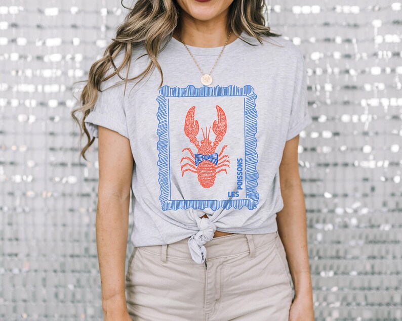 May include: A light gray t-shirt featuring a red lobster design within a blue frame. The lobster has a blue bow tie and the text "LES POISSONS" is printed on the side. The shirt is tied at the waist.