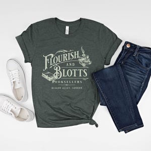 May include: A dark green t-shirt with the text "Flourish and Blotts Booksellers Diagon Alley, London" in white. The shirt is folded and laid out on a white surface with a pair of white sneakers and a pair of blue jeans.