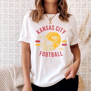 Pode incluir: Camiseta branca com um design gr&aacute;fico vermelho, amarelo e dourado que diz "Kansas City Football Est. 1960" com um gr&aacute;fico de capacete de futebol.