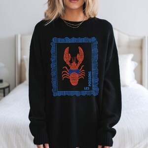 May include: Black sweater featuring a red lobster illustration within a blue frame. The design includes the words "LES POISSONS" in blue. The sweater is a crew neck style.