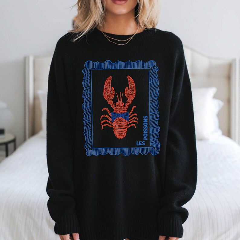 May include: Black sweater featuring a red lobster illustration within a blue frame. The design includes the words "LES POISSONS" in blue. The sweater is a crew neck style.