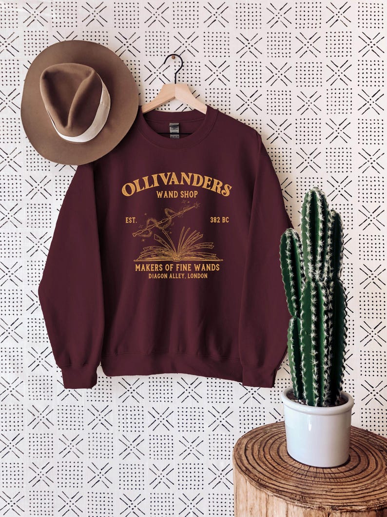 May include: A maroon sweatshirt with a gold graphic of Ollivanders Wand Shop. The graphic includes the text "Ollivanders Wand Shop", "Est. 382 BC", "Makers of Fine Wands", and "Diagon Alley, London".