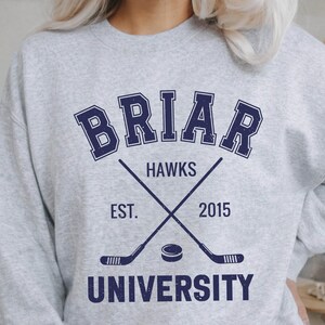May include: A light gray sweatshirt with the text "BRIAR HAWKS UNIVERSITY" in navy blue. The design includes crossed hockey sticks, a hockey puck, and the year "2015". The sweatshirt is a casual apparel item.