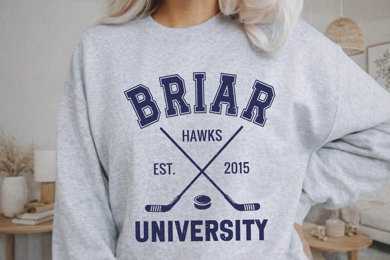 May include: A light gray sweatshirt with the text "BRIAR HAWKS UNIVERSITY" in navy blue. The design includes crossed hockey sticks, a hockey puck, and the year "2015". The sweatshirt is a casual apparel item.