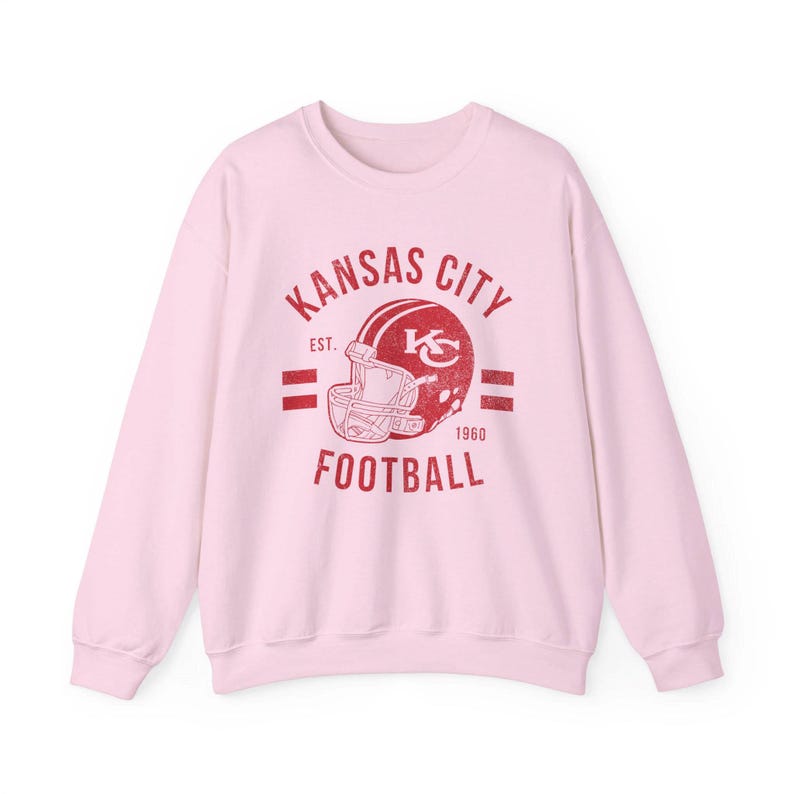 Pode incluir: Camisola cor-de-rosa com um gr&aacute;fico vermelho de um capacete de futebol e o texto "Kansas City Est. 1960 Football"