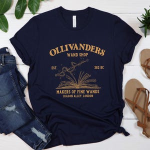 May include: A navy blue t-shirt with a gold graphic of a wand and an open book. The text "Ollivanders Wand Shop Est. 382 BC Makers of Fine Wands Diagon Alley, London" is printed on the shirt.