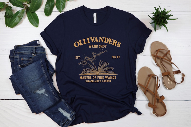 May include: A navy blue t-shirt with a gold graphic of a wand and an open book. The text "Ollivanders Wand Shop Est. 382 BC Makers of Fine Wands Diagon Alley, London" is printed on the shirt.