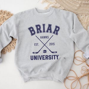 May include: A light gray sweatshirt with the text "BRIAR UNIVERSITY HAWKS EST. 2015" in navy blue. The design includes crossed hockey sticks and a hockey puck. The sweatshirt has a crew neck and long sleeves.