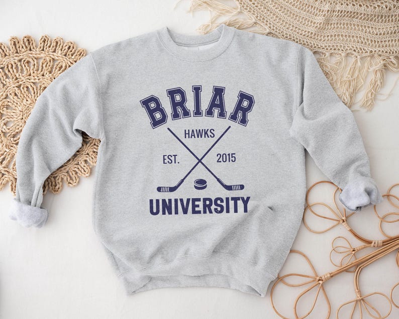 May include: A light gray sweatshirt with the text "BRIAR UNIVERSITY HAWKS EST. 2015" in navy blue. The design includes crossed hockey sticks and a hockey puck. The sweatshirt has a crew neck and long sleeves.