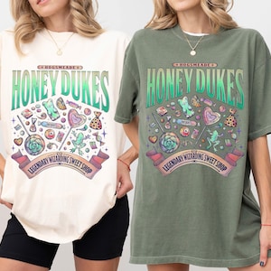 Może przedstawiać: Dwie koszulki, jedna w kolorze kości słoniowej i druga w kolorze mchu, z graficznym wzorem. Wz&oacute;r przedstawia napis "HONEY DUKES" oraz r&oacute;żne słodycze. Obecny jest r&oacute;wnież napis "LEGENDARY WIZARDING SWEET SHOP". Koszulki są z kr&oacute;tkim rękawem.