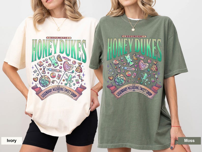 Może przedstawiać: Dwie koszulki, jedna w kolorze kości słoniowej i druga w kolorze mchu, z graficznym wzorem. Wz&oacute;r przedstawia napis "HONEY DUKES" oraz r&oacute;żne słodycze. Obecny jest r&oacute;wnież napis "LEGENDARY WIZARDING SWEET SHOP". Koszulki są z kr&oacute;tkim rękawem.