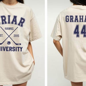 May include: Beige t-shirt with navy blue text and graphics. The front features "Briar Hawks University" with crossed hockey sticks and a puck. The back displays "Graham 44". A casual, oversized fit.