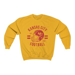 Pode incluir: Uma camisa de moletom amarela com um logotipo vermelho e branco do Kansas City Football. O logotipo apresenta um capacete de futebol com as letras "KC" dentro. O texto "Kansas City" est&aacute; acima do capacete e "Football" est&aacute; abaixo. O texto "Est. 1960" est&aacute; &agrave; direita do capacete.