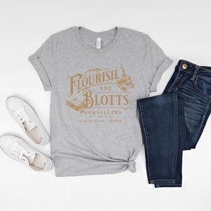 May include: A gray t-shirt with the text "Flourish and Blotts Booksellers Diagon Alley, London" in gold lettering. The shirt is paired with blue jeans and white sneakers.