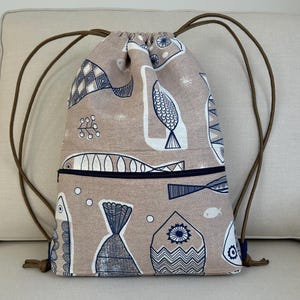 May include: A beige drawstring backpack featuring a repeating pattern of stylized blue and white fish and aquatic shapes. The bag has a black zip pocket and brown drawstring cords. The bag is made of fabric.