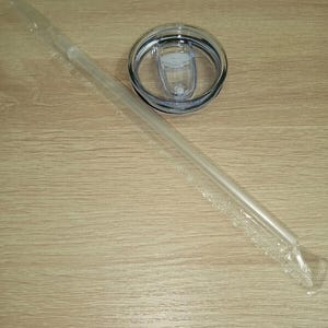 Pu&ograve; includere: Una cannuccia di plastica trasparente in un involucro di plastica, un coperchio trasparente con chiusura scorrevole e un bordo nero. Il coperchio &egrave; progettato per un bicchiere o una tazza da viaggio. Gli oggetti sono su una superficie di legno marrone chiaro.