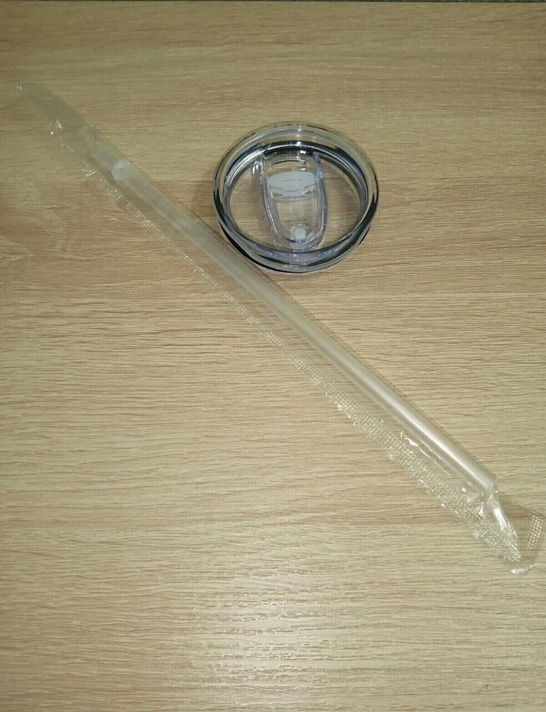 Pu&ograve; includere: Una cannuccia di plastica trasparente in un involucro di plastica, un coperchio trasparente con chiusura scorrevole e un bordo nero. Il coperchio &egrave; progettato per un bicchiere o una tazza da viaggio. Gli oggetti sono su una superficie di legno marrone chiaro.