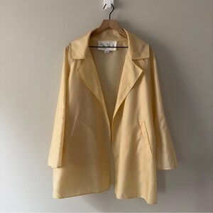 May include: A pale yellow open-front jacket with a collar and two front pockets hangs on a wooden hanger. The jacket is made of a smooth, lightweight fabric. The jacket is hanging against a neutral wall.