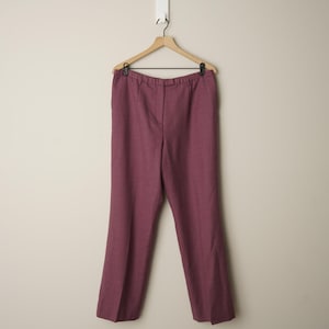 Puede incluir: Pantalones de color burdeos colgados de una percha de madera. Los pantalones de pernera recta tienen un diseño clásico y están hechos de un material tejido. Los pantalones están suspendidos sobre un fondo neutro. La longitud de los pantalones es de unos 100 cm.