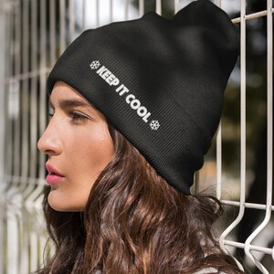 May include: A black beanie with the text "KEEP IT COOL" in white, along with snowflake graphics. The beanie is worn by a person with long, wavy brown hair and a gray top. The background features a white metal fence.