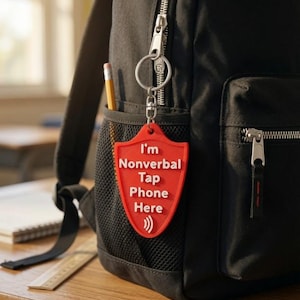 May include: A red shield-shaped keyring attached to a black backpack. The keyring features white embossed text that reads: I'm Nonverbal Tap Phone Here, accompanied by a white wireless signal icon at the bottom of the shield-shaped surface.