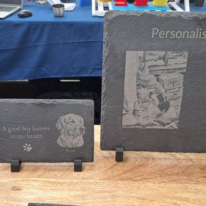 May include: Three rectangular dark grey slate plaques with rough edges displayed on stands. The left plaque shows a dog, the centre plaque features a dog with the text A good boy forever in our hearts, and the right plaque displays a photo and text.