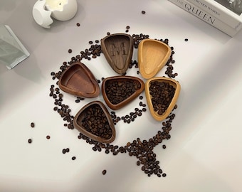 May include: Six wooden, triangular-shaped bowls, some filled with coffee beans, arranged on a white surface. The bowls vary in color from light to dark brown. Coffee beans are scattered around the bowls. One bowl has the initials "M T" carved into it.