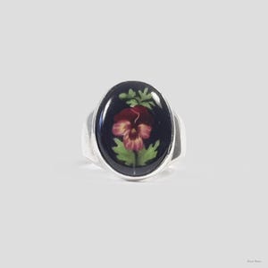 May include: A silver ring with a black oval setting featuring a pressed flower design of a purple pansy.