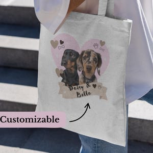 Peut inclure: Un sac fourre-tout en toile blanche avec un motif de cœur rose représentant deux chiens. Les chiens sont un teckel noir et fauve et un teckel bringé. Le texte "Daisy & Bella" est imprimé sur une bannière brune sous les chiens.