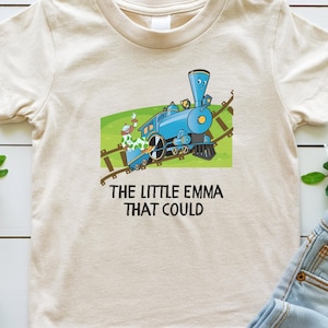 May include: A beige t-shirt with a cartoon illustration of a blue train on a green background. The text "THE LITTLE EMMA THAT COULD" is printed below the image.