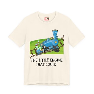 May include: Cream-colored t-shirt featuring a blue cartoon train on a green field with brown train tracks. The text "THE LITTLE ENGINE THAT COULD" is printed below the train. The shirt has short sleeves and a crew neck.