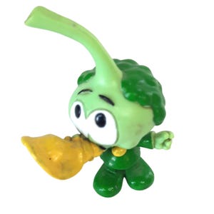 May include: A green broccoli character figurine with a yellow horn. The figurine has a cartoonish design with large eyes and a smiling expression. The broccoli head is a darker green, and the stem is a lighter green.