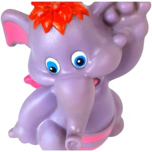Könnte beinhalten: Eine lila Elefantenfigur mit großen blauen Augen, rosa Ohren und einer roten blütenartigen Verzierung auf dem Kopf. Der Elefant hat ein rosa-weiß gestreiftes Band um seinen Bauch. Das Spielzeug besteht aus einem glatten, glänzenden Material.