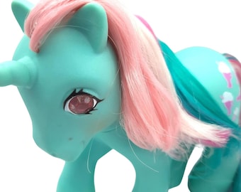 May include: A turquoise unicorn toy with pink and white hair. The unicorn has a white horn, pink eyes, and a design of ice cream cones on its flank. The toy is a vintage collectible.