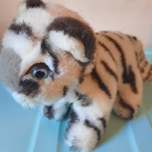 May include: A plush tiger cub toy with a tan body and black stripes. The toy has a white and brown head with large blue eyes and a small mouth. The toy is standing on a light blue surface.