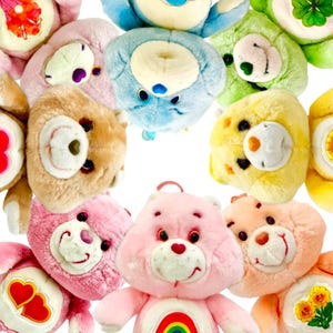 May include: A collection of plush bear toys in various colors including pink, blue, yellow, green, and brown. Each bear features a distinct embroidered symbol on its belly, such as a rainbow, hearts, flowers, and a clover, with soft fur and stitched facial features.