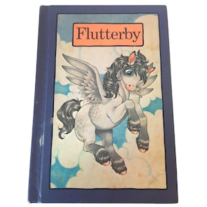 May include: A hardcover book titled "Flutterby" with a blue cover. The cover illustration features a winged horse with a gray body, dark gray mane and tail, and pink accents. The background shows a blue sky with white clouds.