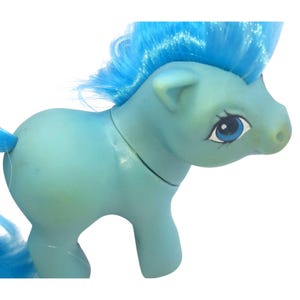 May include: A light blue toy pony with a blue mane and tail. The pony has a detailed face with blue eyes and a white outline. The toy is made of plastic and is a vintage collectible.