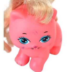 Op de afbeelding: Een roze kattenbeeldje met blauwe ogen en blond haar. De speelgoedkat heeft een lachend gezicht met zwarte snorharen en wenkbrauwen. De kat is een vintage speelgoed met wat slijtage.