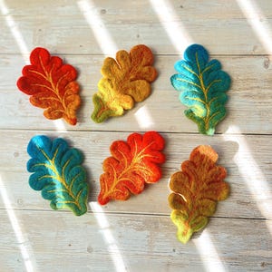 May include: Six felt oak leaves in various shades of orange, yellow, and blue. The leaves are arranged in a semi-circle on a light wood surface.