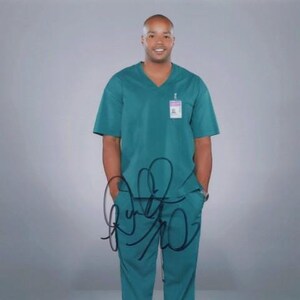 May include: A man wearing a teal scrub top with a name tag and black pants. He is standing in front of a gray background and has a signature on the bottom of the image.
