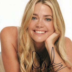 May include: A portrait of a smiling woman with long, blonde hair and blue eyes, resting her chin on her hand. The image has a white background. An autograph is visible on the arm.
