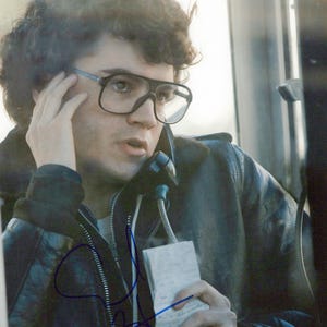 May include: A man wearing large black-framed glasses and a black leather jacket is on the phone in a phone booth. He is holding a notepad and the phone receiver. The image is a vintage photograph.
