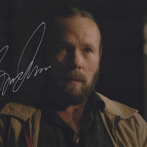 May include: A signed photograph featuring a man with a beard, wearing a tan jacket over a dark shirt. The image has the text "THE LAST OF US" in the lower left corner and a signature in the upper left corner.