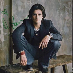 Puede incluir: Un hombre con un blazer azul oscuro con un collar de cadena de plata y una pulsera de plata se sienta en un banco de madera. Está mirando a la cámara. El fondo es una pared gris.