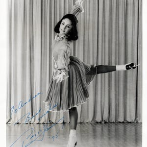 Może przedstawiać: Czarno-białe zdjęcie łyżwiarki figurowej Peggy Fleming w pozie podczas występu. Ma na sobie plisowaną sukienkę, łyżwy i ozdobę głowy. Obraz zawiera autograf i tekst "Peggy Fleming '75".