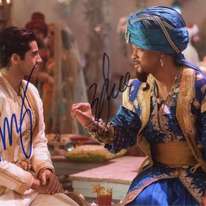 May include: Two men in elaborate costumes, one wearing a blue turban and the other a white and gold robe, stand in a room with ornate decorations. The man in the turban is holding his hand up in a gesture of greeting or blessing. The man in the robe is looking at the man in the turban.