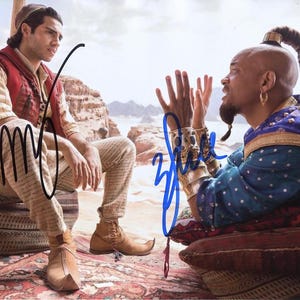 May include: A signed photo of two actors, one wearing a blue and gold costume and the other wearing a red and white costume. The actor in the blue and gold costume is holding his hands up in the air, while the actor in the red and white costume is sitting on a brown cushion. The photo is signed "Will" and "Mena".