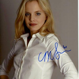 May include: A woman with long blonde hair wearing a white button-down shirt. She is looking at the camera and has a slight smile on her face. The shirt is slightly sheer and her signature is visible on the right side of the image.